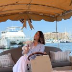 Woman holding a puppy on a boat with a marina and large yacht in background, under a canopy with striped pillows.