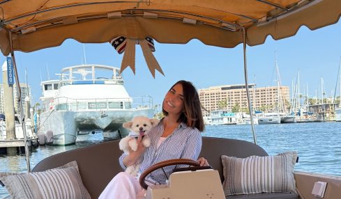 Woman holding a puppy on a boat with a marina and large yacht in background, under a canopy with striped pillows.