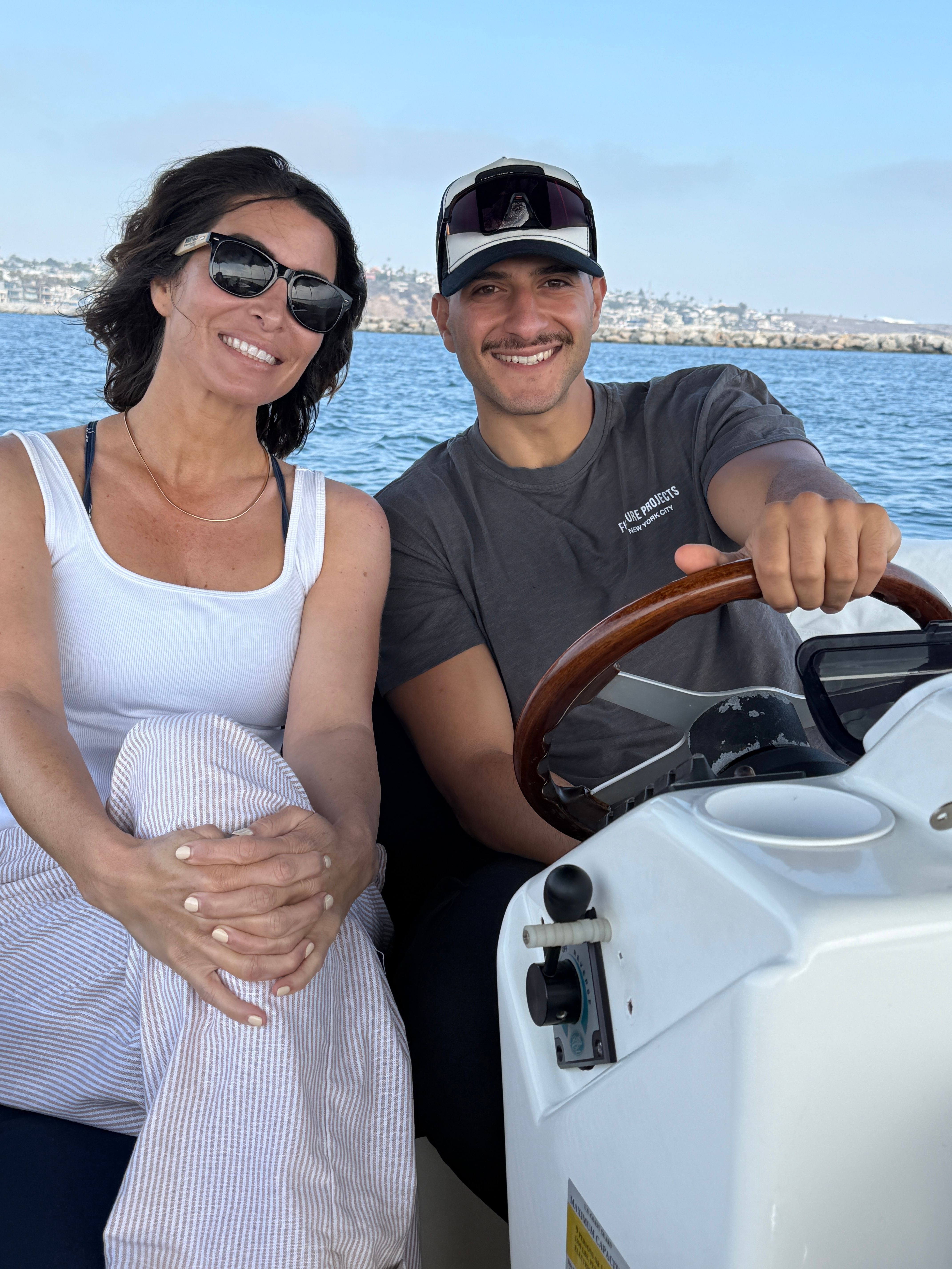 A woman and a man are on a boat, smiling, with the woman holding the man's hand. The woman wears sunglasses and a white top. The man wears a cap and a dark shirt, steering the boat. Water and sky are visible in the background.