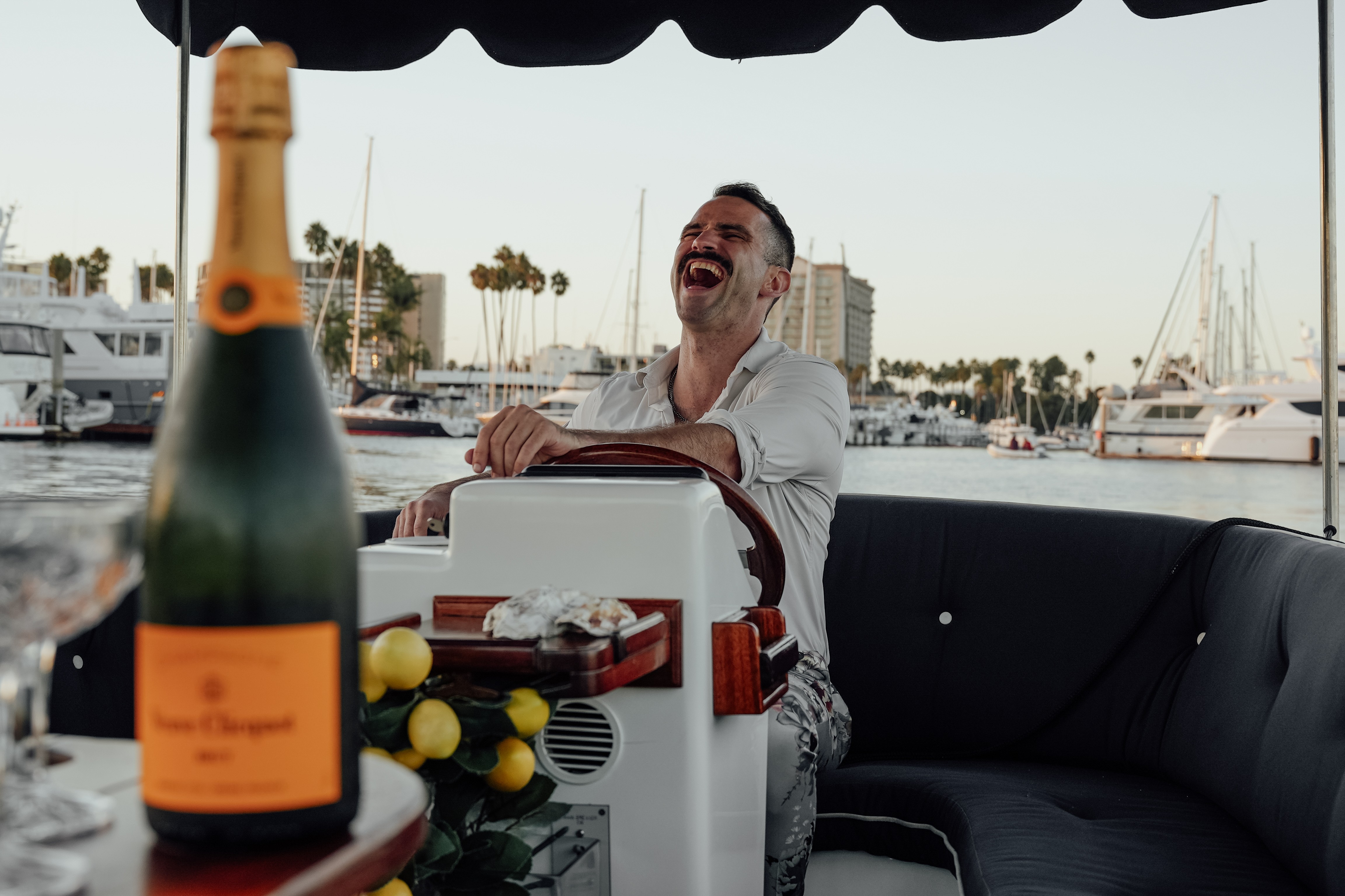 Man laughing on a boat with a marina and yachts in the background, a champagne bottle in foreground, and a black sofa.
