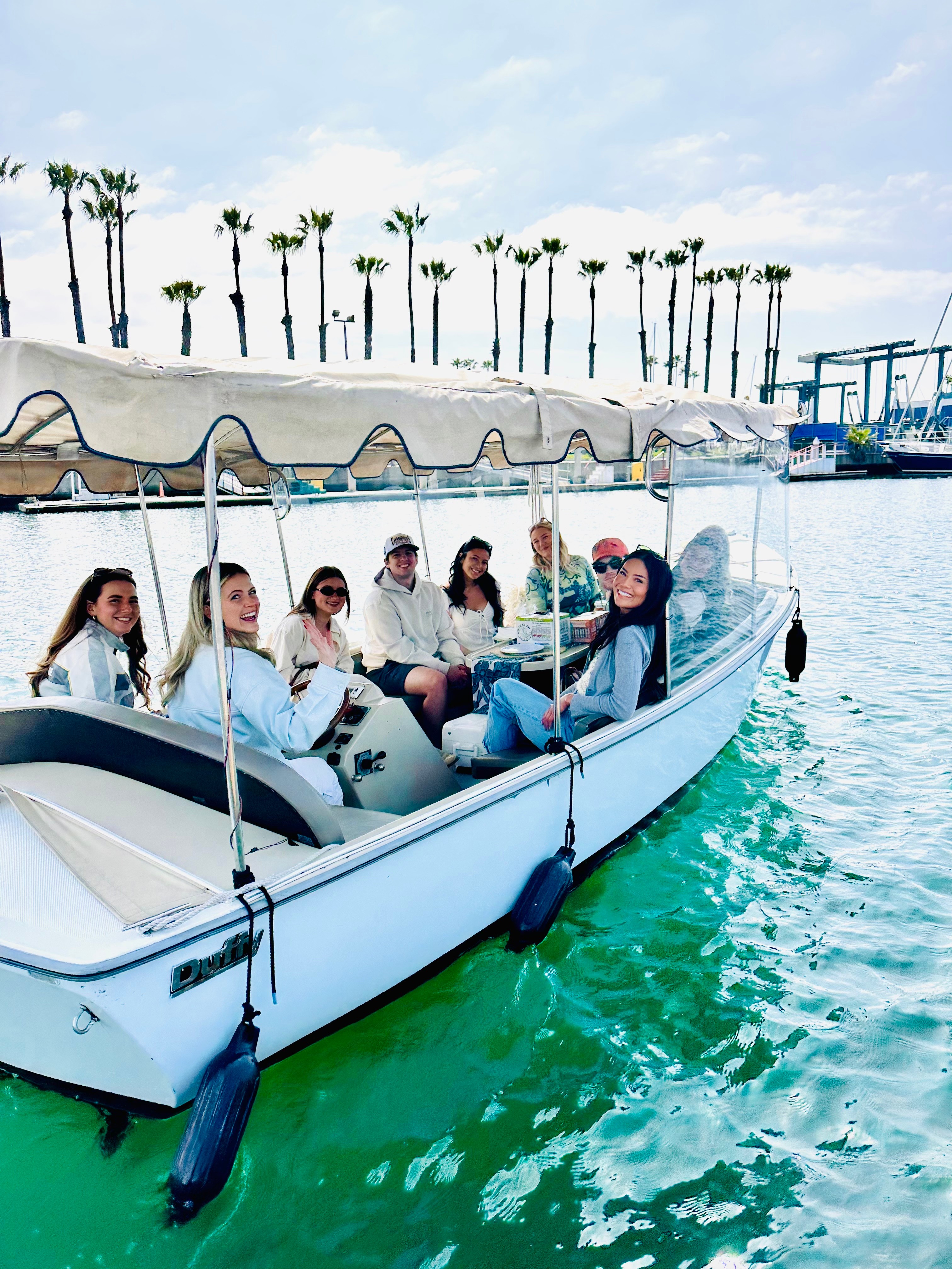 Group of people sitting on a boat with a canopy, on water with palm trees and a marina in background.