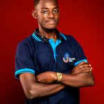 Young man with arms crossed, wearing a navy blue polo shirt with blue accents, standing against a red background.