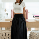 Woman standing indoors near a desk, smiling, with large window and trees outside in background.