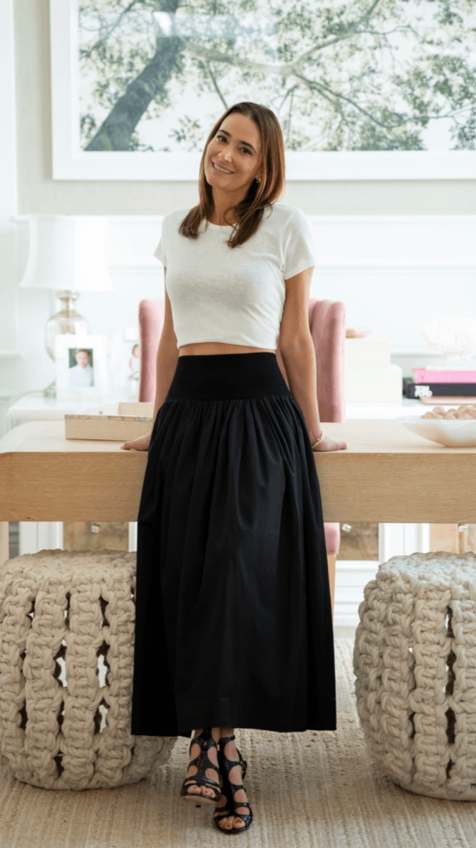 Woman standing indoors near a desk, smiling, with large window and trees outside in background.