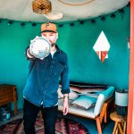 Person wearing a cap and denim jacket pointing at the camera in a room with teal walls and a bed.