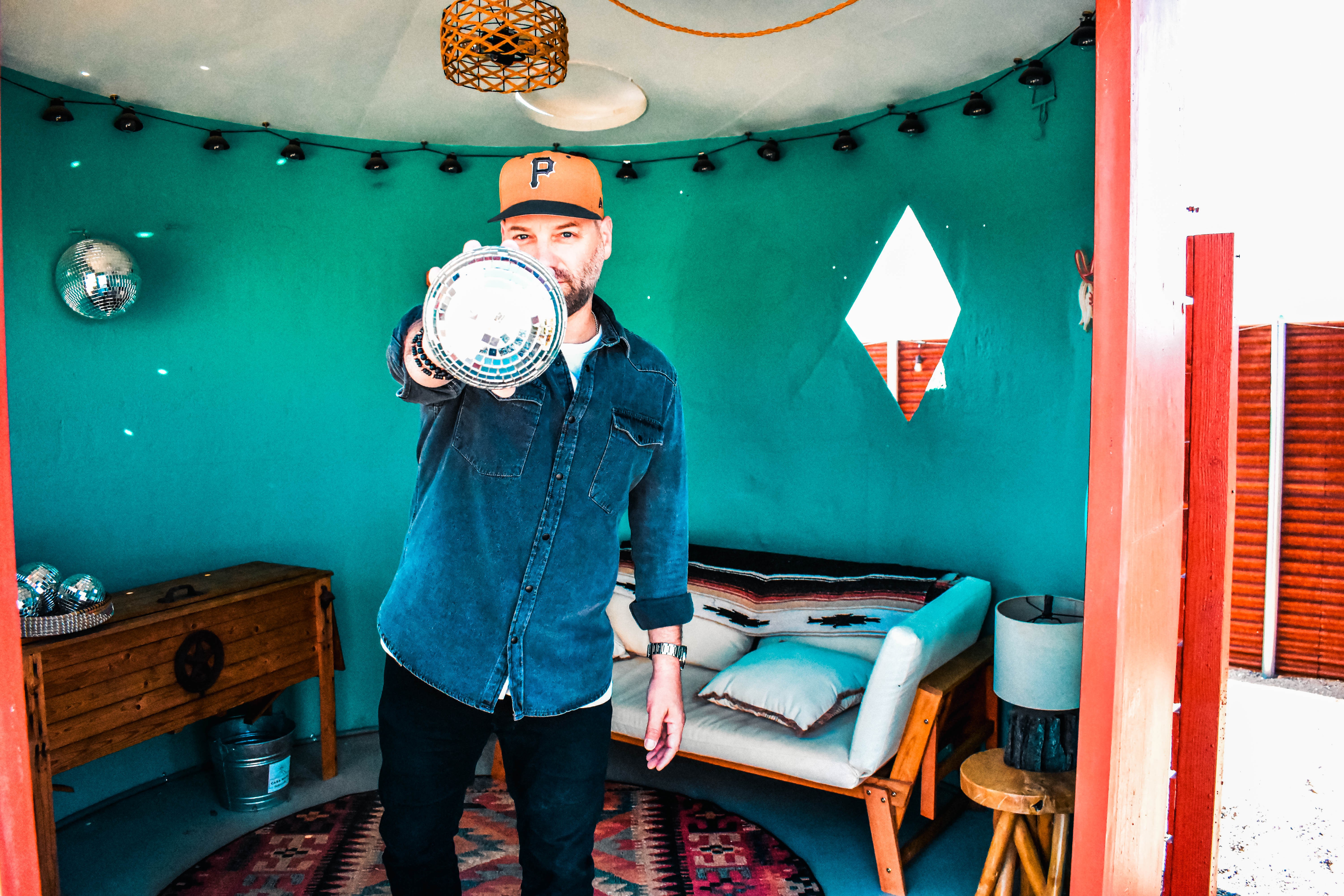 Person wearing a cap and denim jacket pointing at the camera in a room with teal walls and a bed.