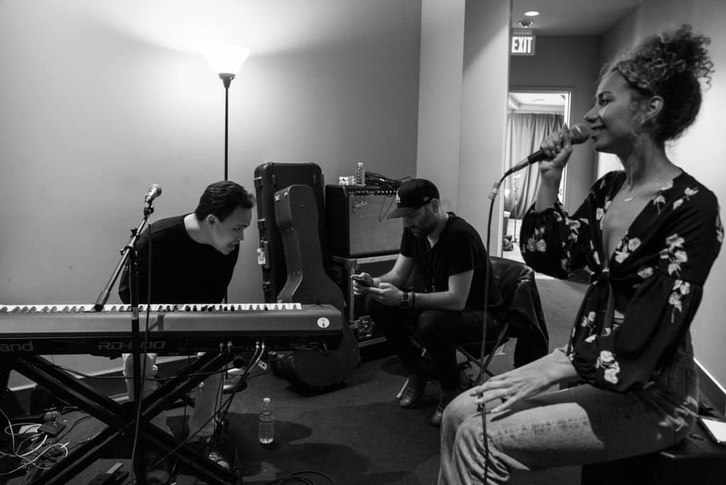 Three musicians in a room: a woman singing into a microphone, two men playing keyboard and guitar, with musical equipment around.