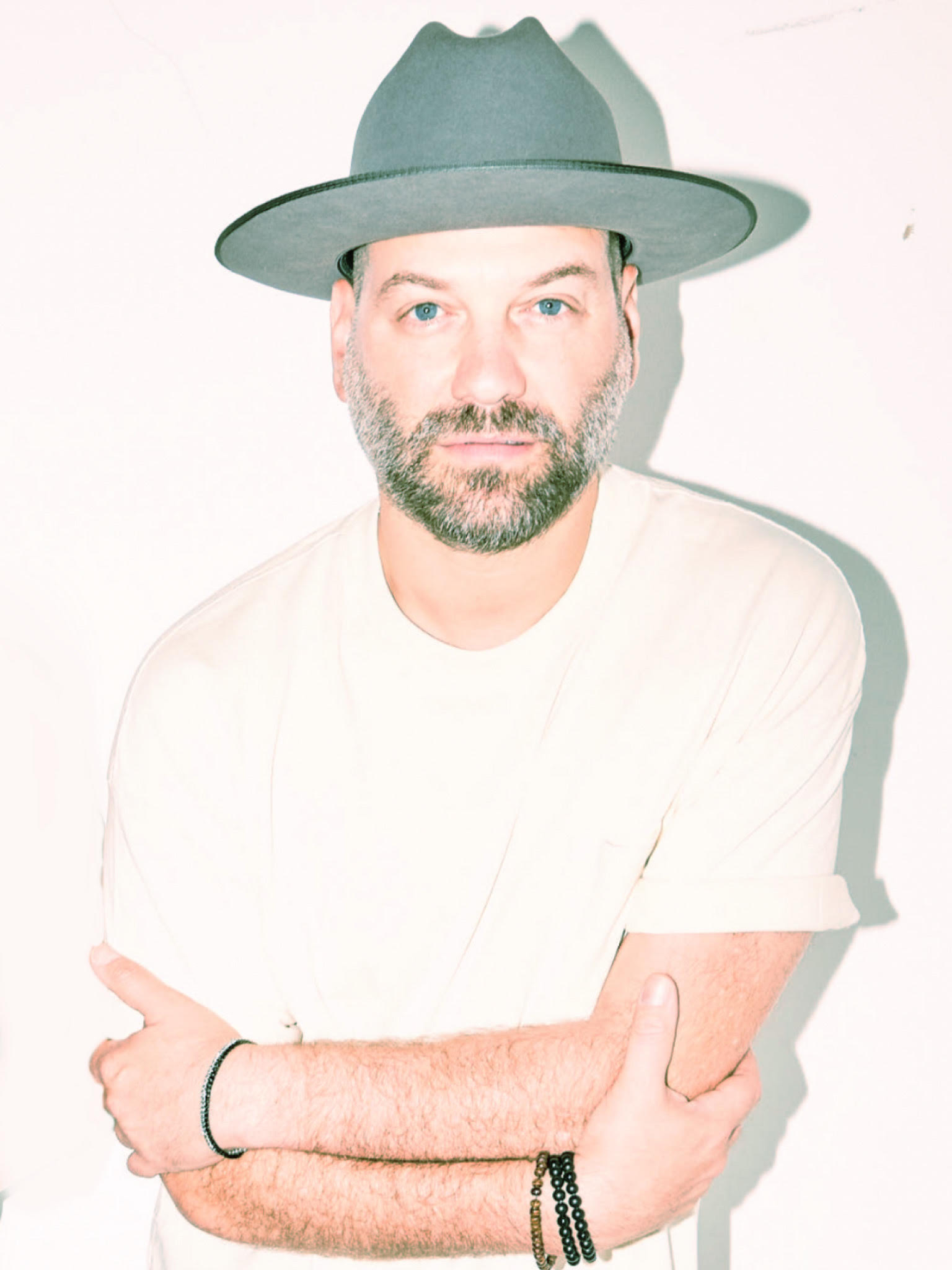 Man with a beard wearing a wide-brimmed hat and white shirt, crossing arms, looking at camera, shadow cast on white background.