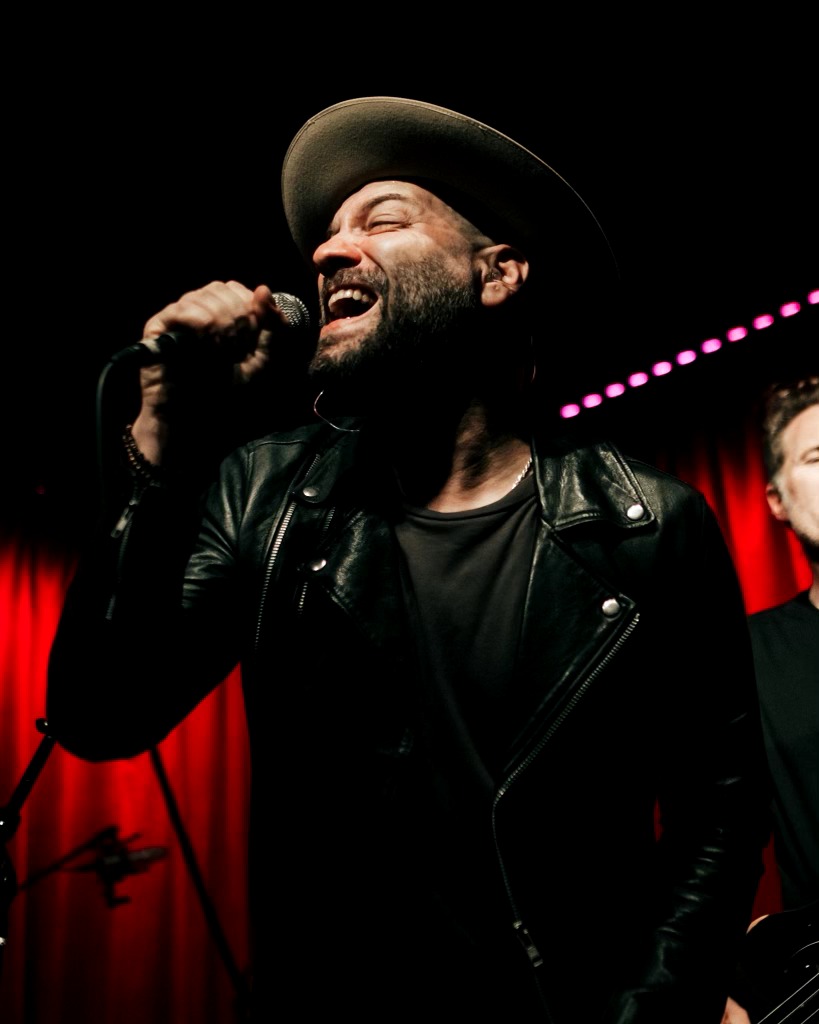 Man singing into a microphone, wearing a wide-brimmed hat and leather jacket, with a red curtain background.