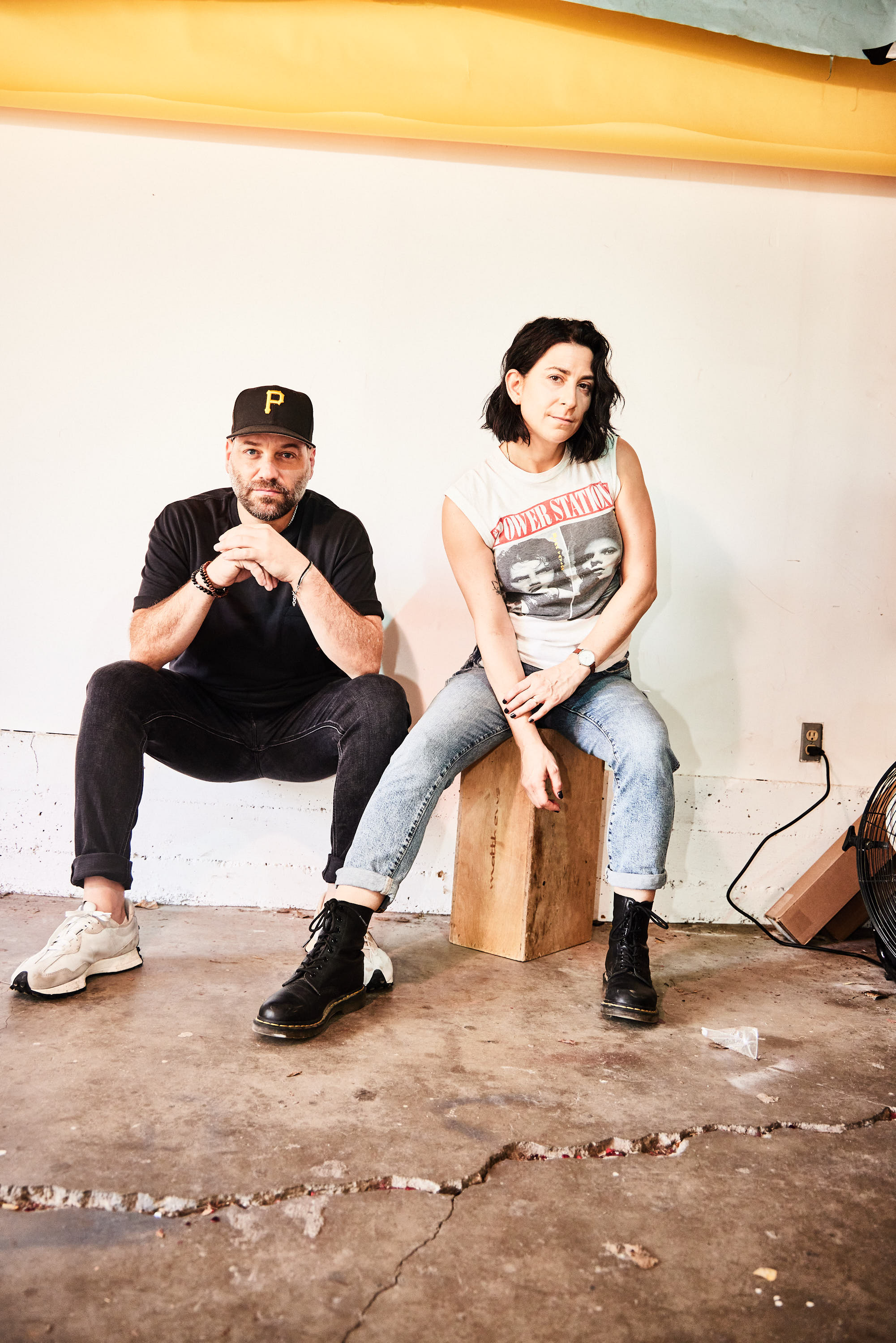 Two people sitting on a wooden block against a plain white wall, one with a black cap and the other with a graphic t-shirt.