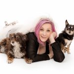 Woman with pink hair lying on her stomach with two dogs, one fluffy and one small, on a white background.