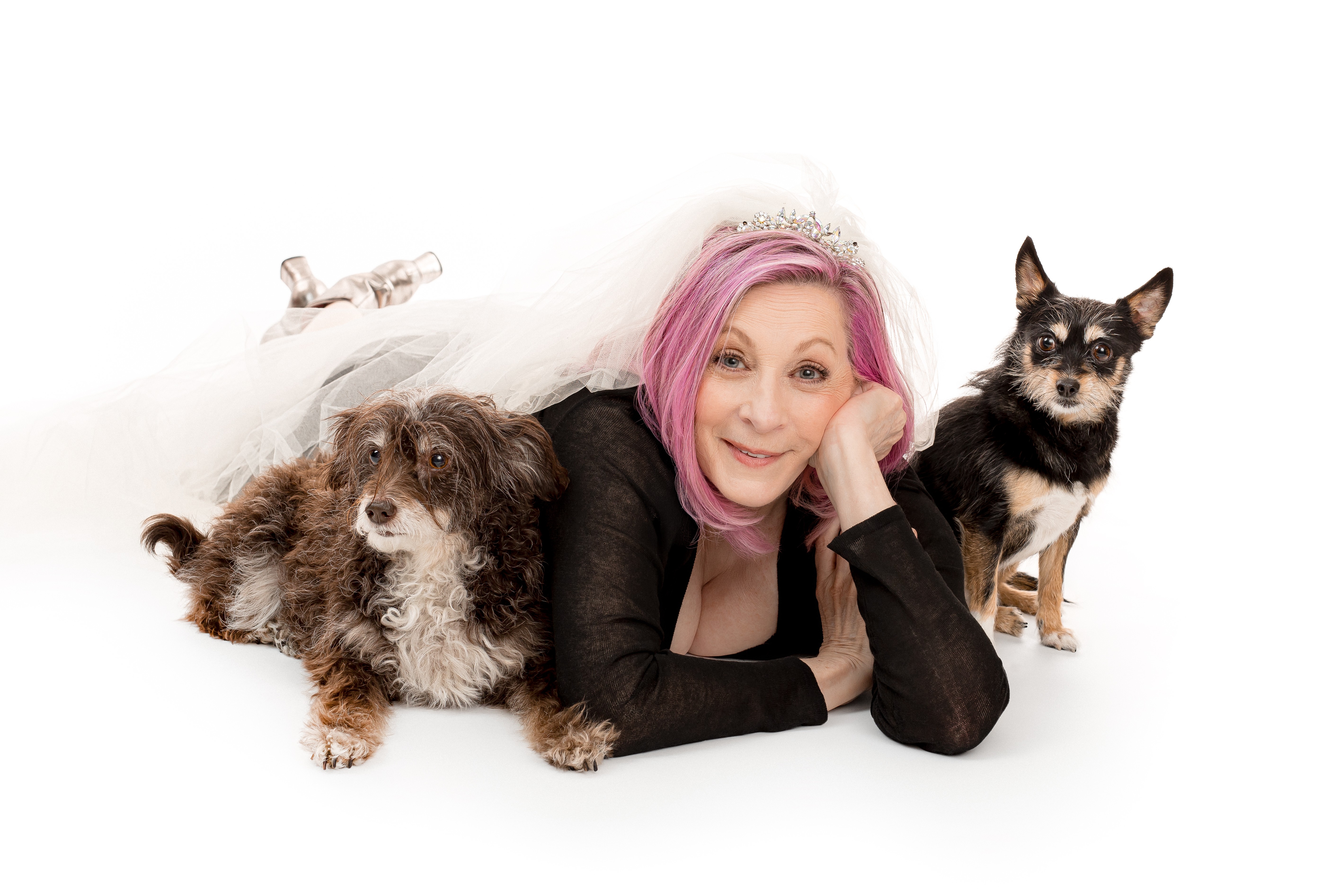 Woman with pink hair lying on her stomach with two dogs, one fluffy and one small, on a white background.