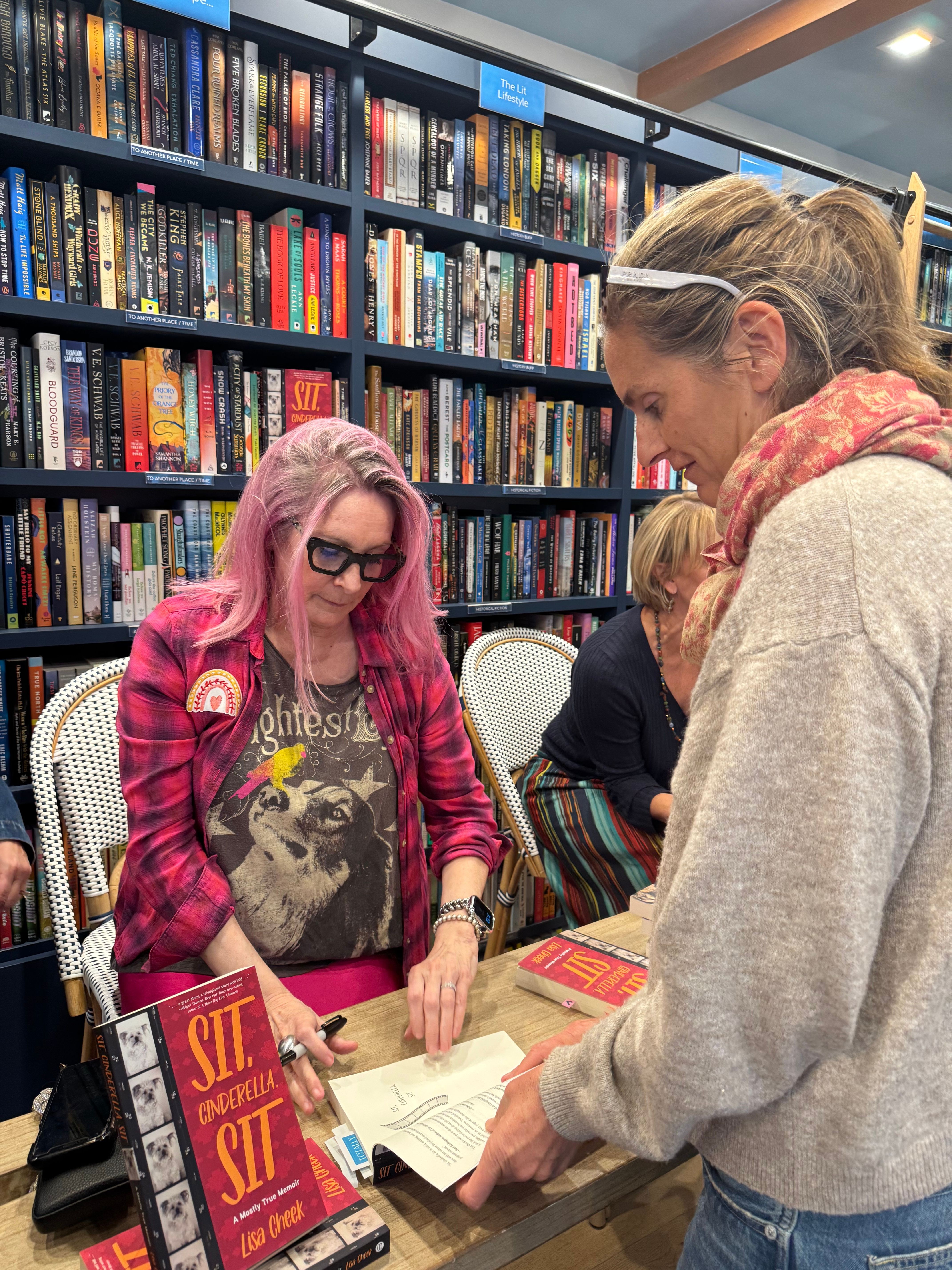 Two women sit at a table in a bookstore, browsing books with shelves filled with books in the background.