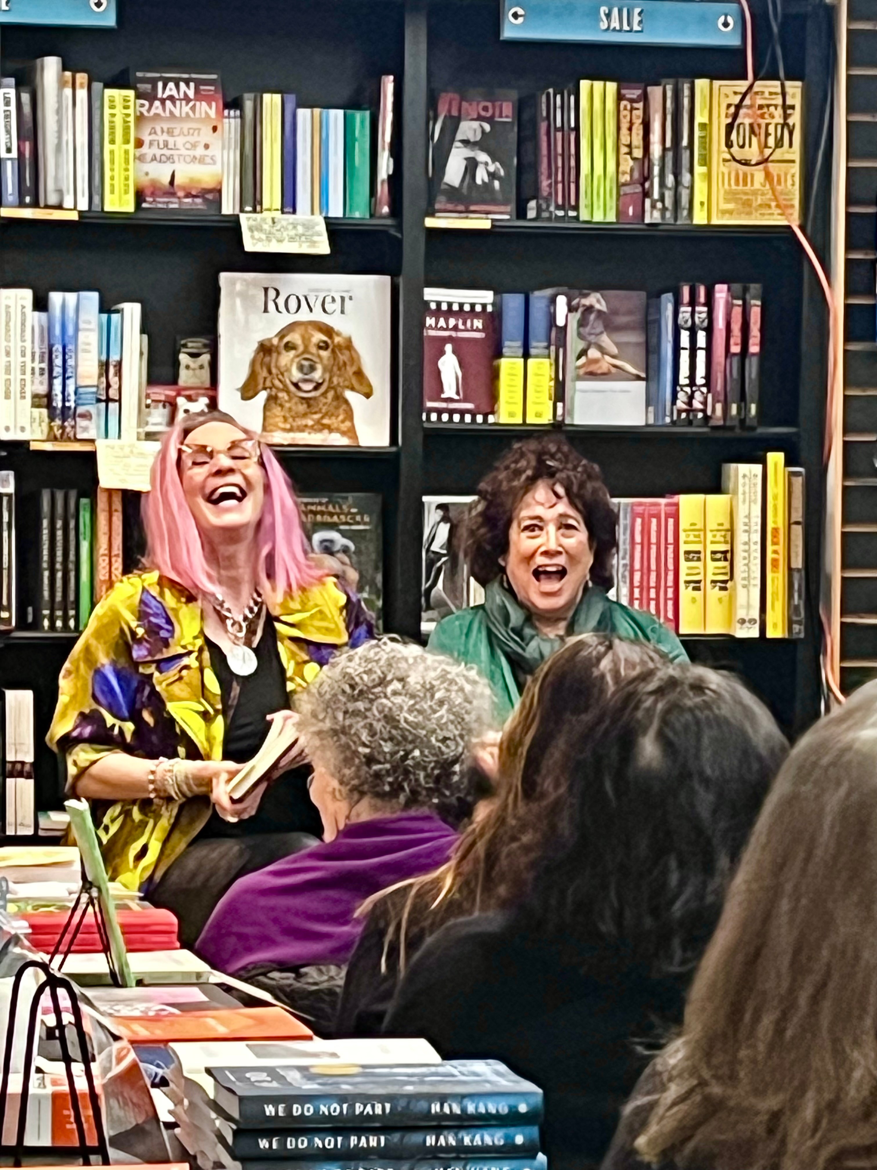 Two women laughing and speaking to an audience in a bookstore with shelves of books behind them.
