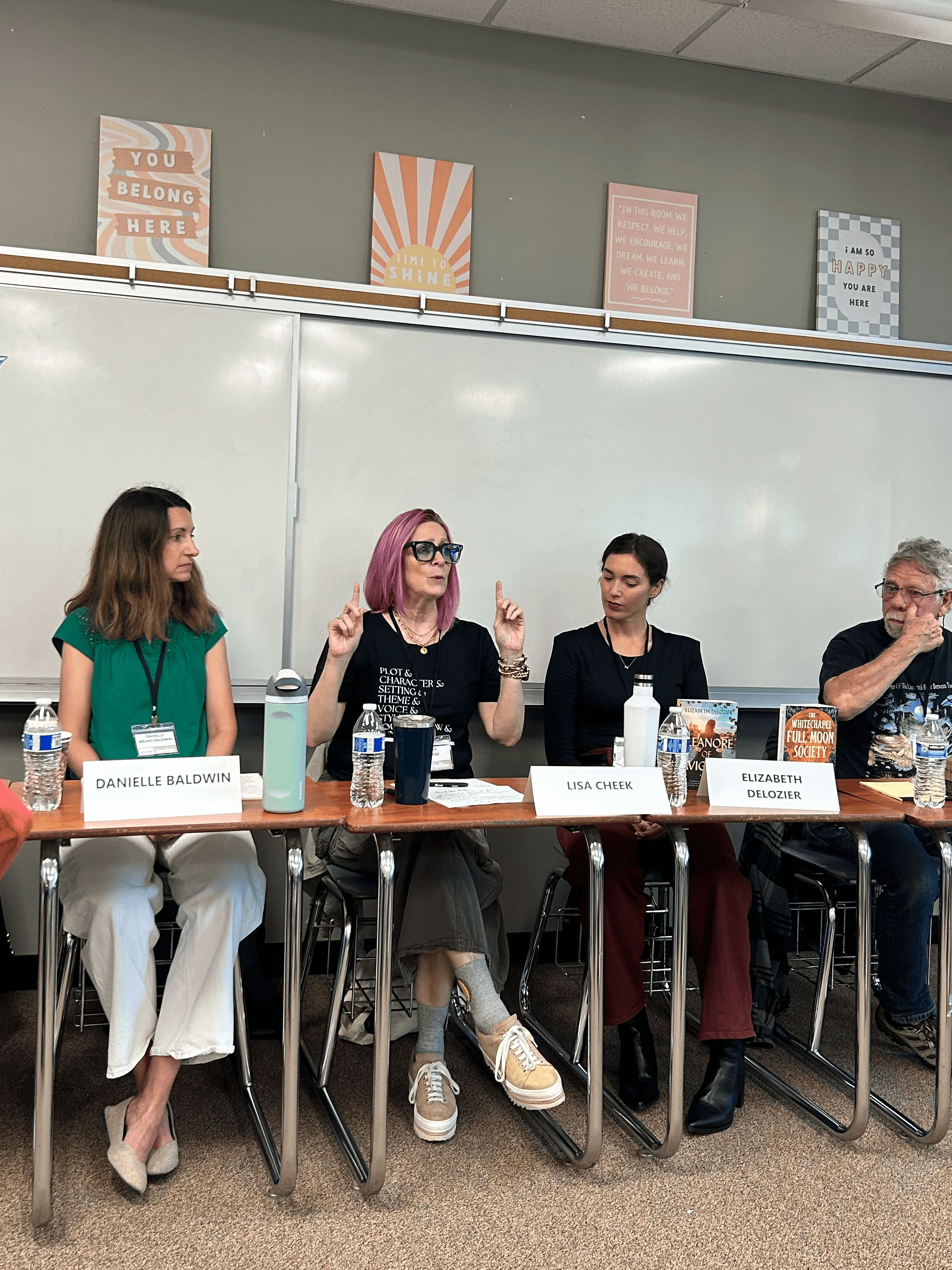 Four people seated at a panel discussion in a classroom, with a whiteboard and colorful posters behind them.