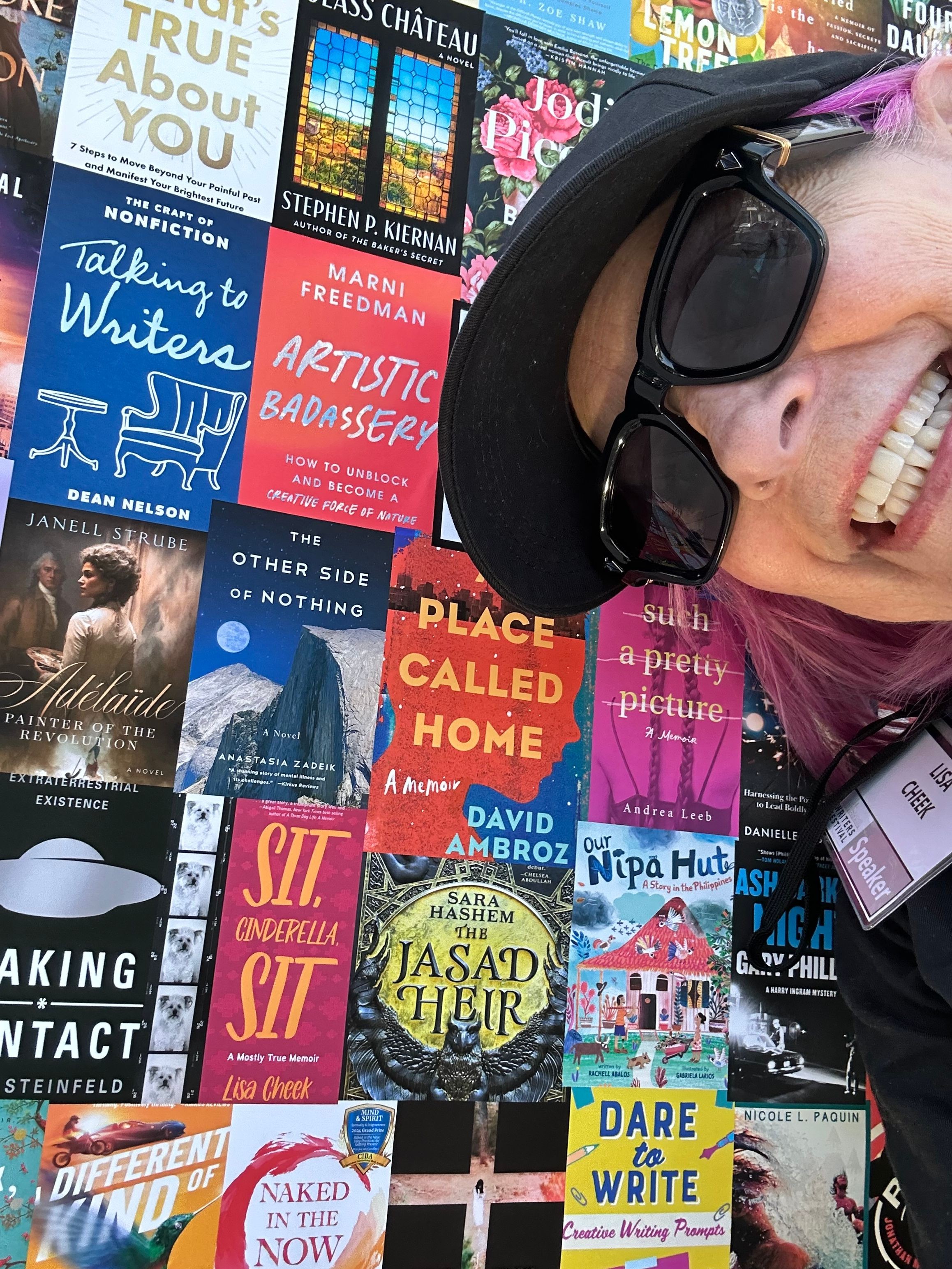 Person with pink hair and sunglasses smiling, surrounded by colorful books and magazines on a table.