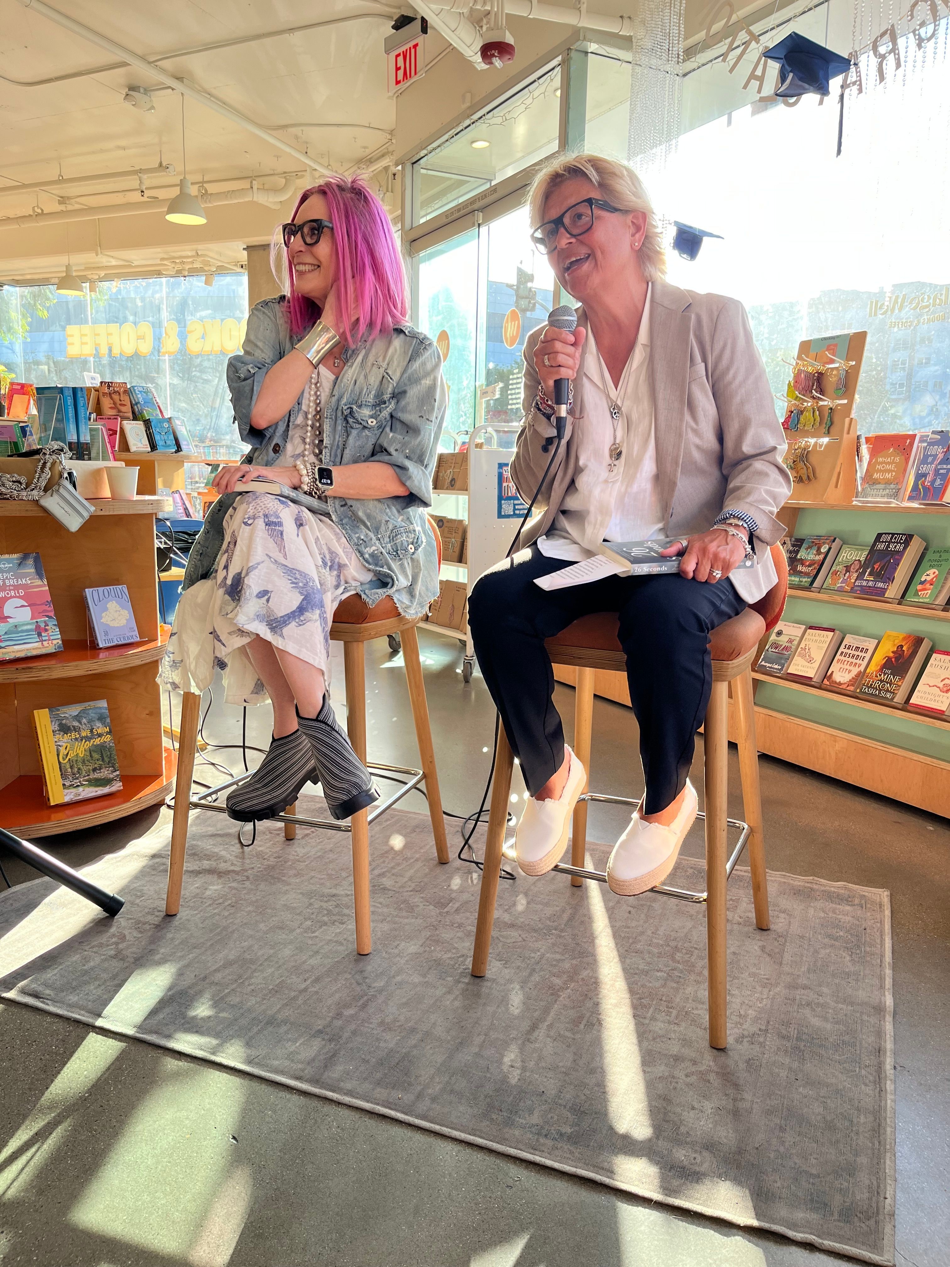 Two women sitting on chairs in a bookstore, one with blonde hair and glasses, the other with pink hair and boots.