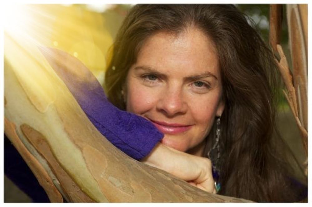 Woman with long hair smiling, resting her head on a tree branch in sunlight.