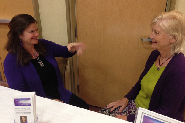 Two women sitting and talking in a room, smiling at each other, with a table and framed photos in front of them.