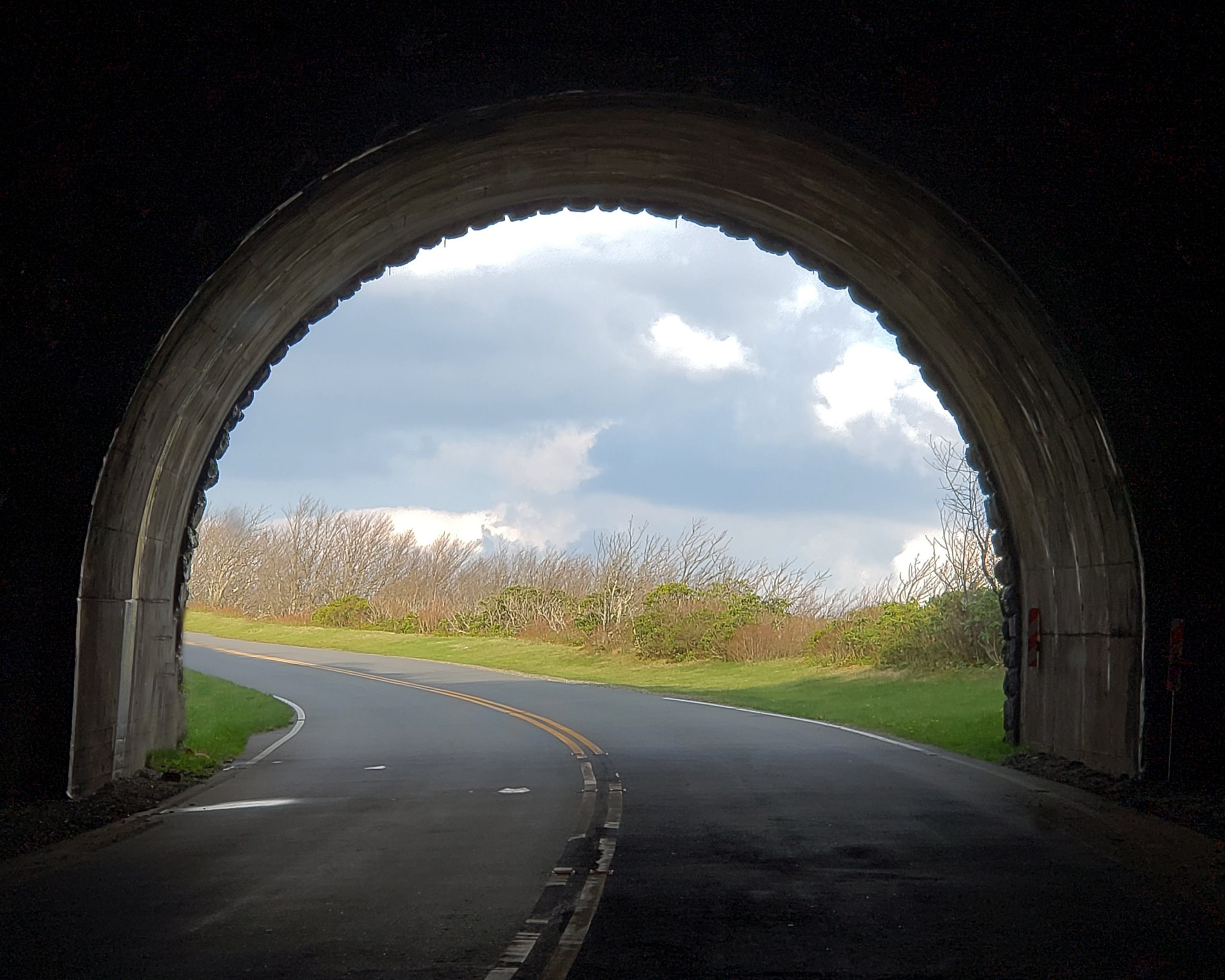 View from inside a tunnel looking out at a curved road and landscape with trees and cloudy sky.