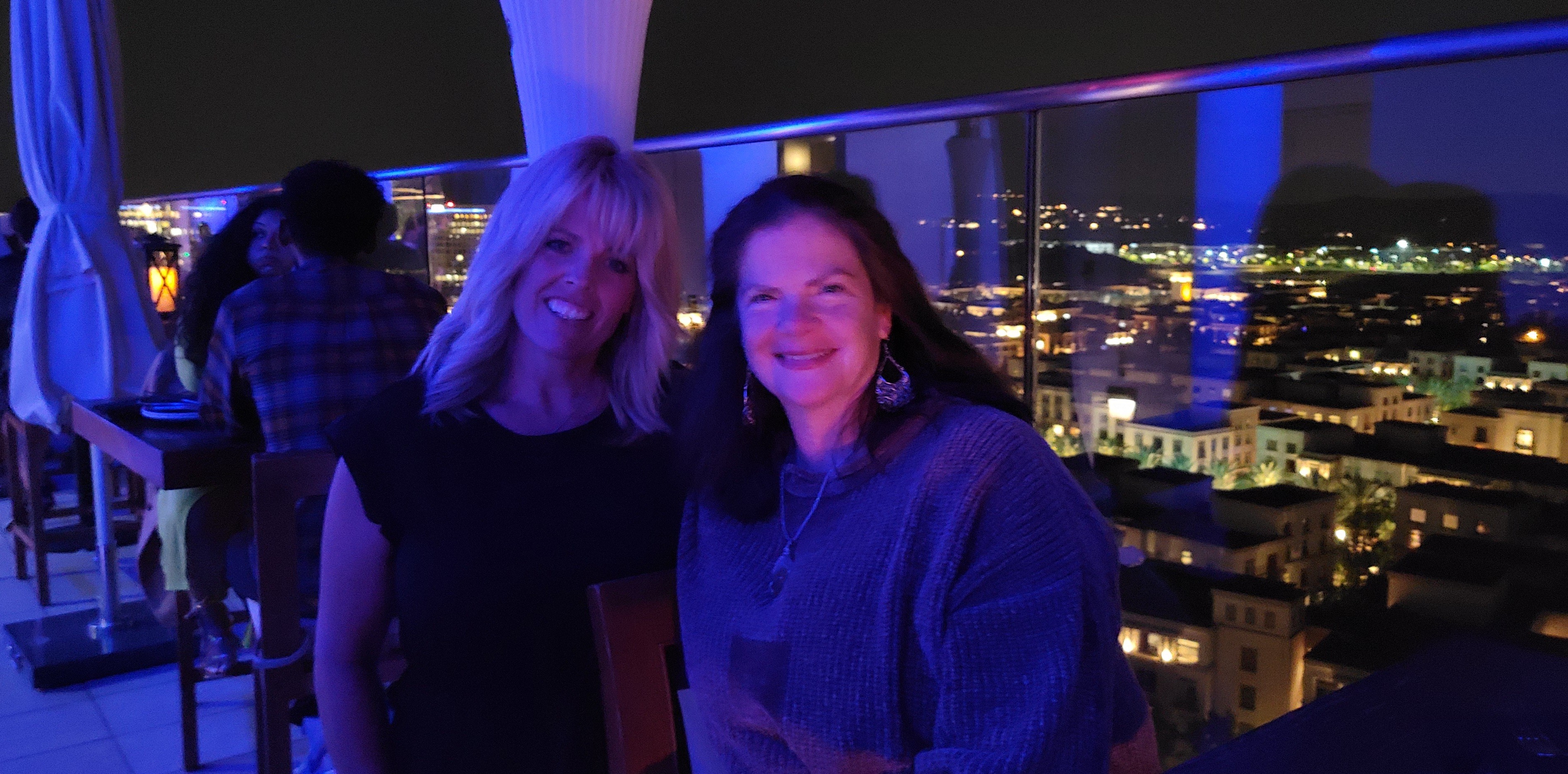 Two women smiling on a balcony at night with city lights in the background.