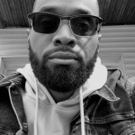Man with sunglasses and beard wearing hoodie and denim jacket, looking at camera, outdoor setting, black and white.