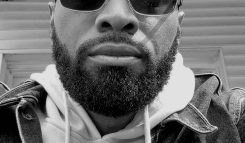 Man with sunglasses and beard wearing hoodie and denim jacket, looking at camera, outdoor setting, black and white.