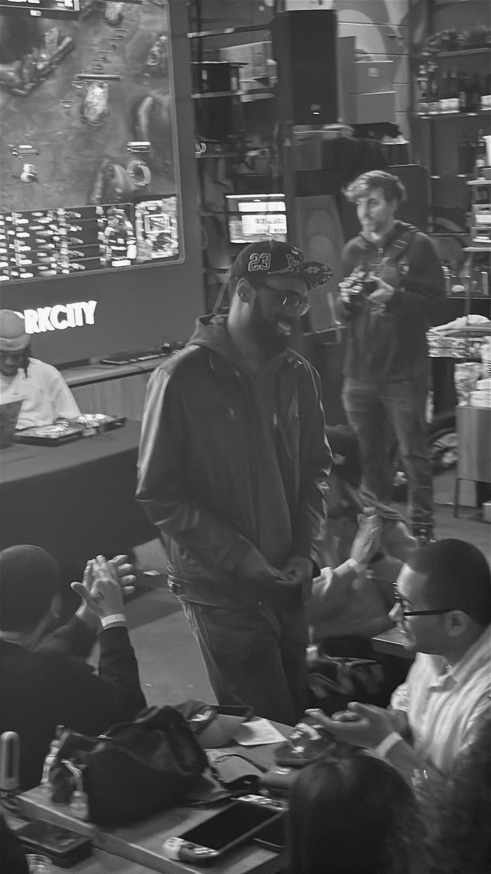 People gathered in a gaming or entertainment venue with screens and gaming equipment, some seated and some standing, in black and white.