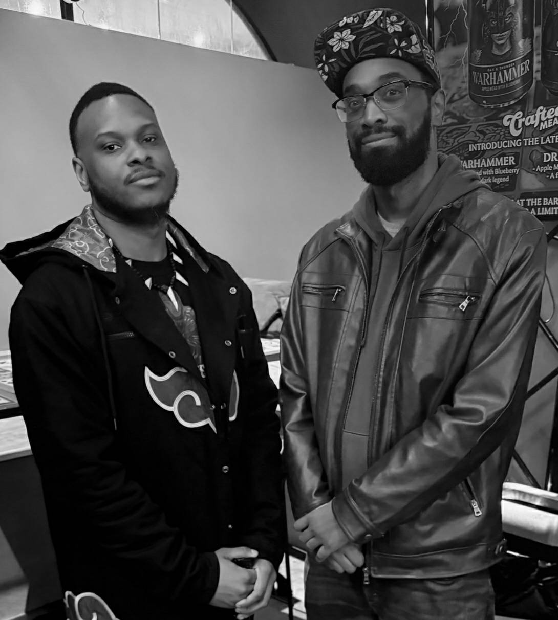 Two men standing indoors, one wearing a hoodie and the other a leather jacket and cap, in front of a plain background.