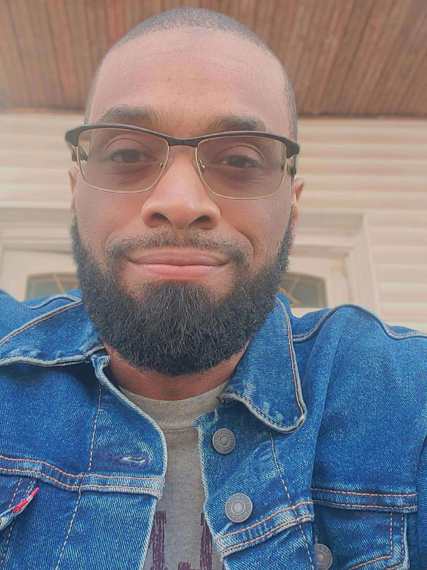 Man with glasses and beard smiling outdoors, wearing a denim jacket, background with wooden ceiling and siding.