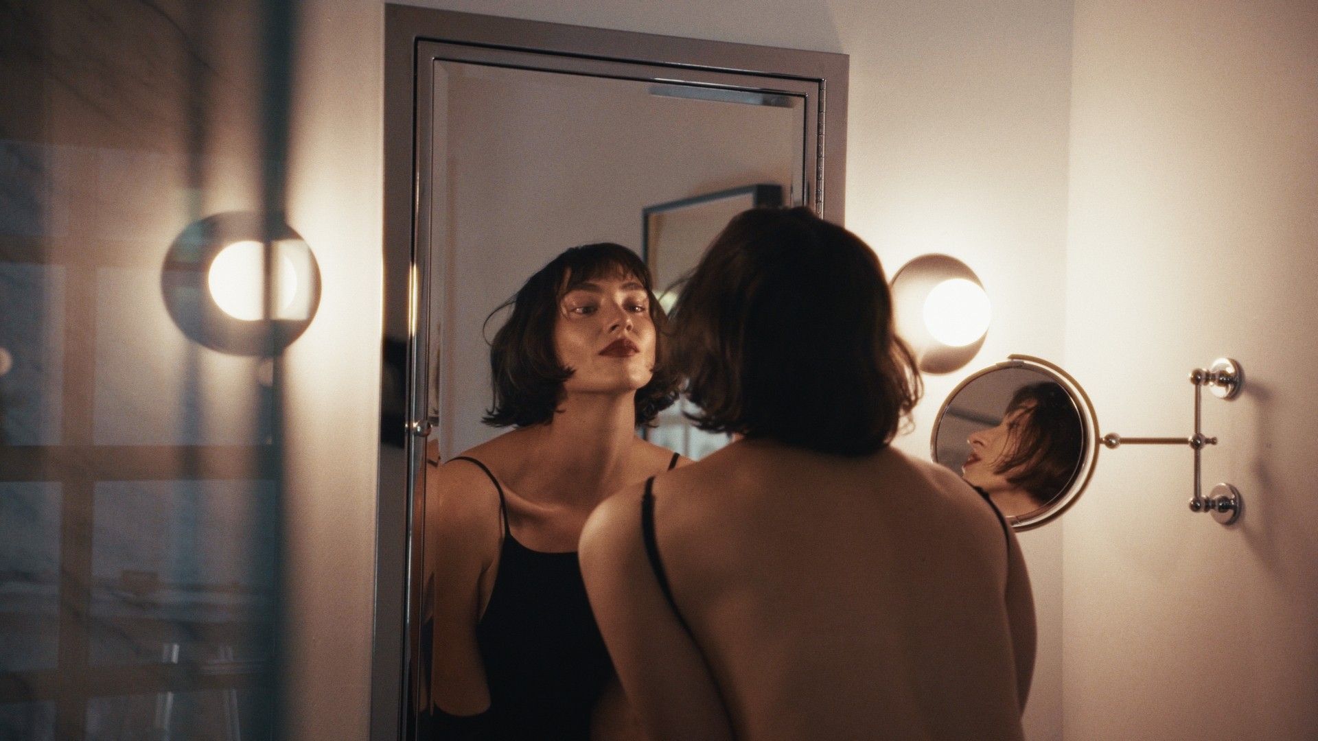 Woman with short dark hair looking at her reflection in a mirror, illuminated by wall-mounted lights, in a dimly lit room.
