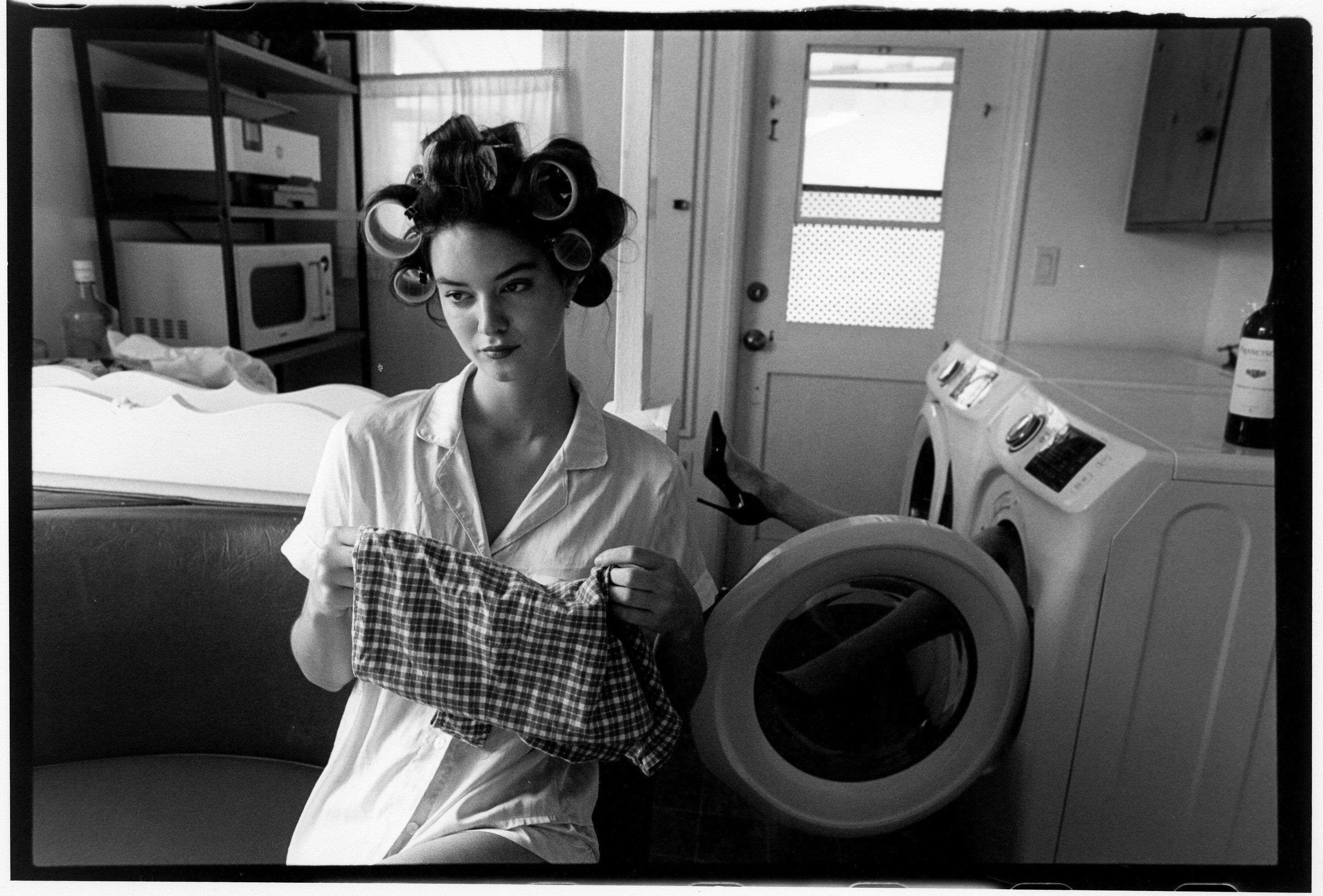 Woman with hair rollers holding a checkered cloth in a laundry room, washing machine nearby, black and white photo.
