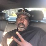 Man with a beard and mustache wearing a black cap and black shirt, sitting in a car, making a peace sign.