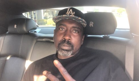 Man with a beard and mustache wearing a black cap and black shirt, sitting in a car, making a peace sign.