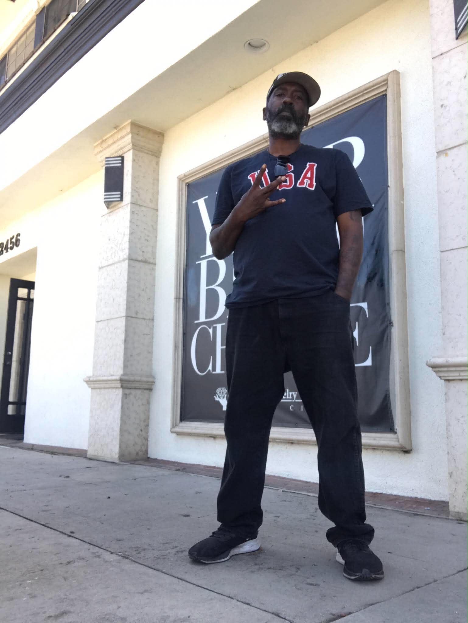 Man standing on sidewalk in front of building, wearing a dark T-shirt with USA, making a peace sign, smiling.