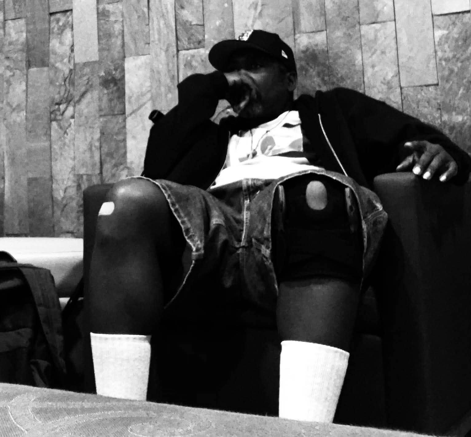 Person sitting in a chair with legs spread, wearing a cap, jacket, shorts, and socks, against a wooden wall background.