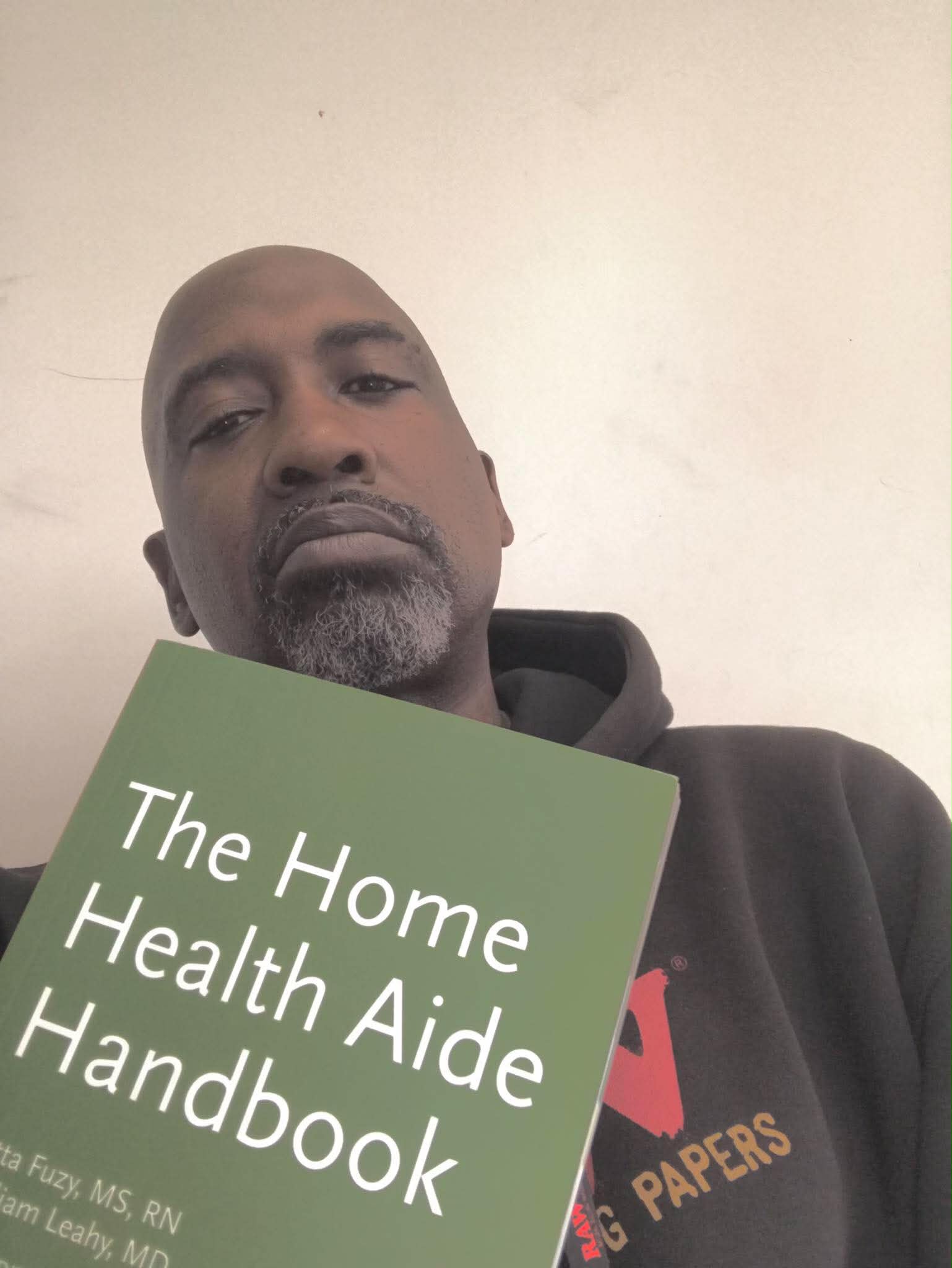 Man holding a green book titled 'The Home Health Aide Handbook' with a plain background.