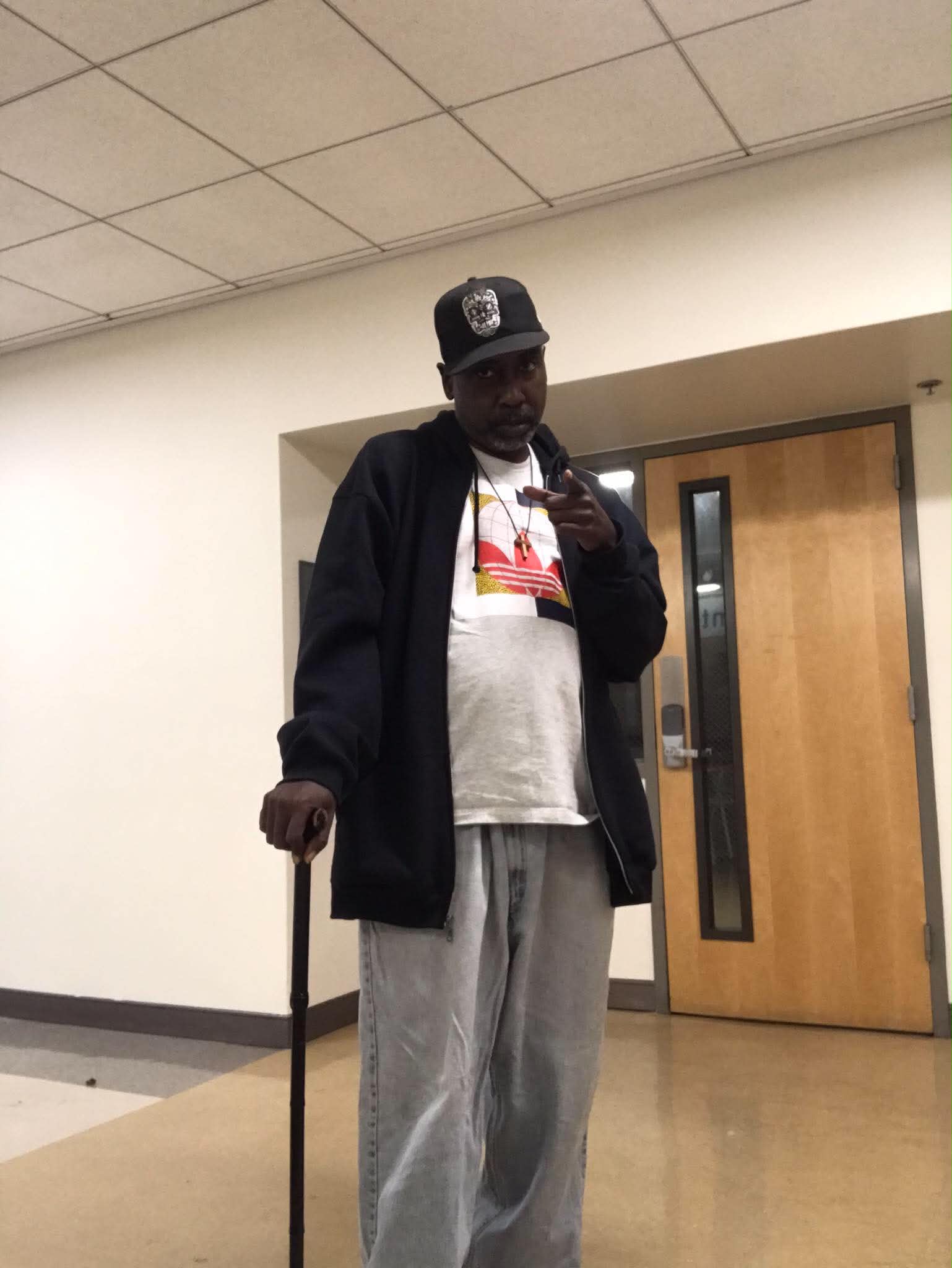 Person standing indoors, wearing a cap, jacket, and gray pants, holding a cane, and making a peace sign gesture.
