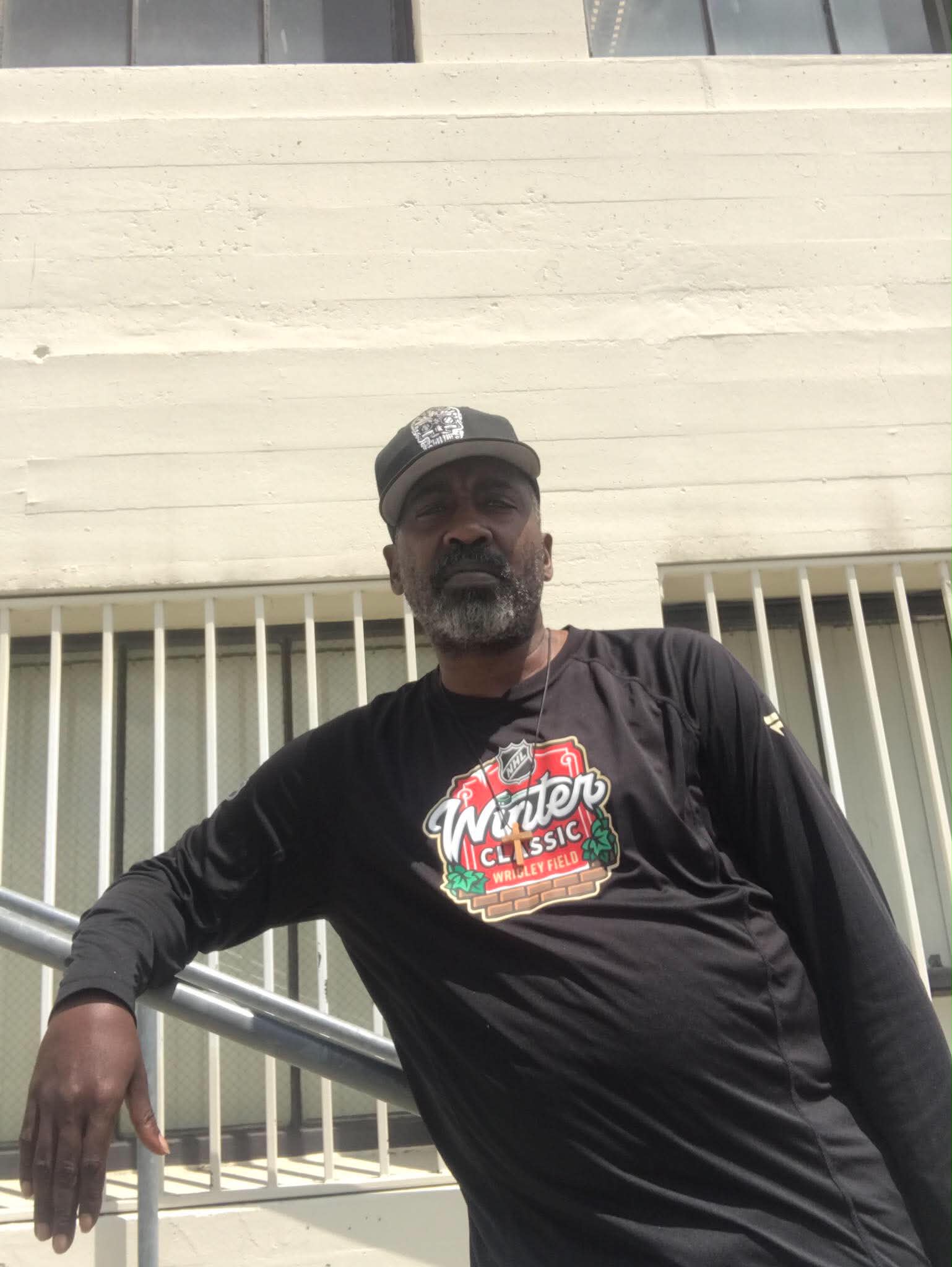 Man with a beard wearing a black cap and black T-shirt leaning on a railing outside, with a building in the background.