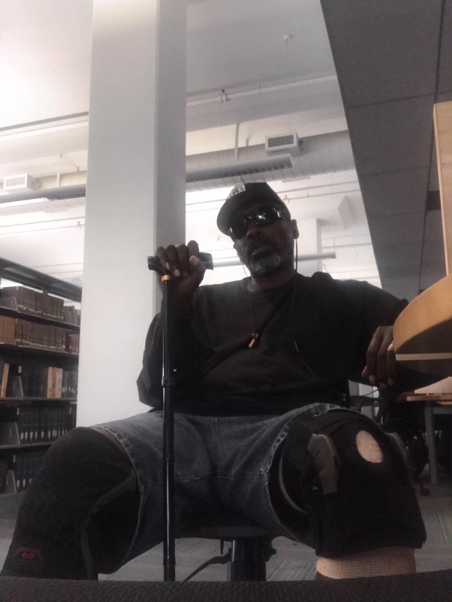 Person sitting in a room with bookshelves, wearing sunglasses, holding a cane, and dressed in dark clothing.