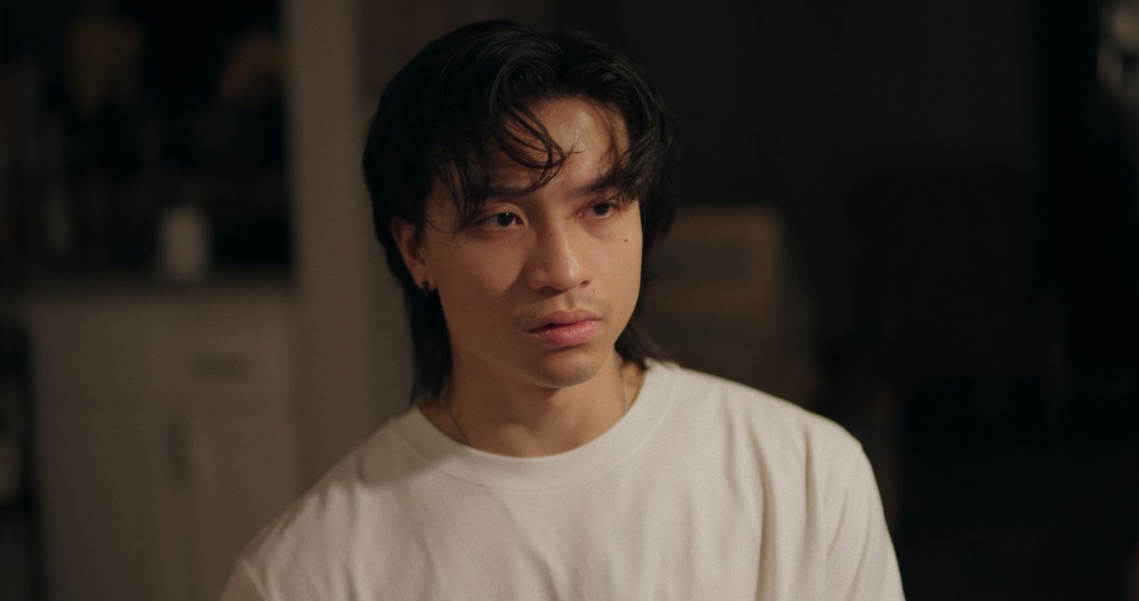 Young person with dark hair wearing a white shirt, looking to the side in a dimly lit room.
