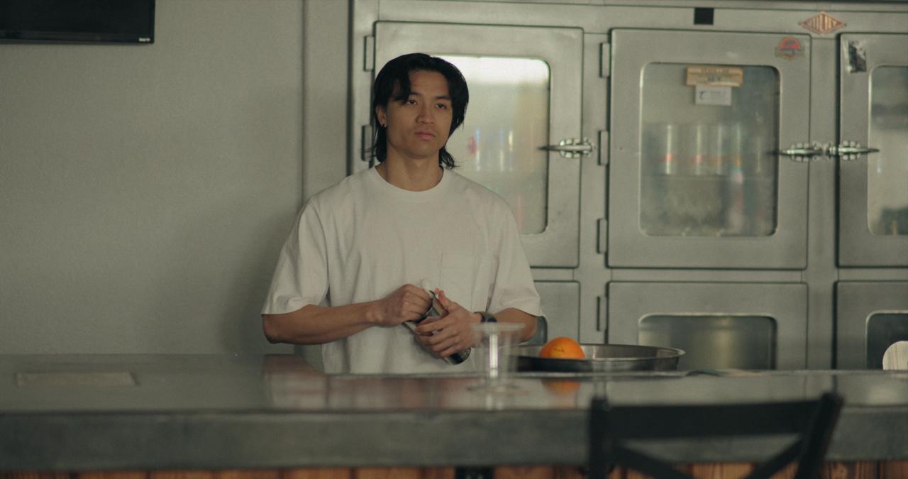 Person standing in a kitchen with stainless steel appliances, wearing a white shirt, hands clasped, looking to the side.