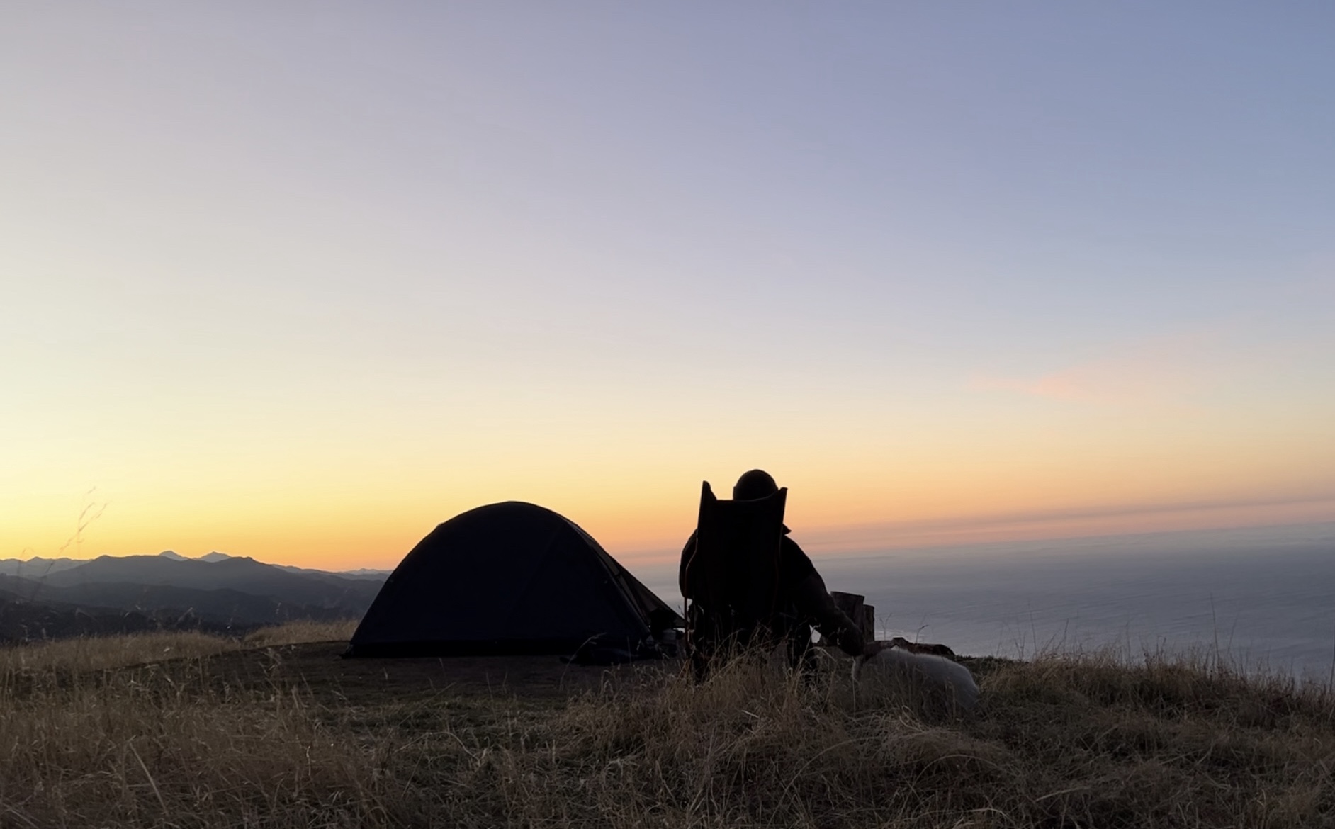 Person sitting near a tent on a grassy hill during sunset, with a view of the ocean in the background.