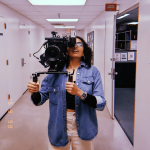 Person holding a camera in a hallway, wearing glasses and a denim jacket, taking a selfie.