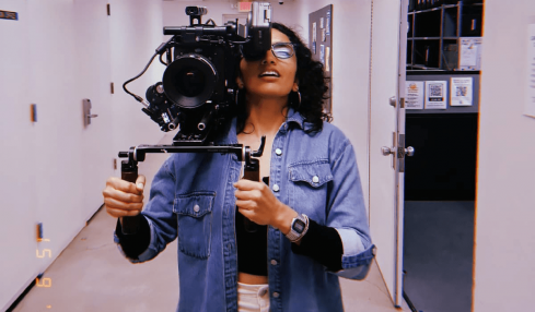 Person holding a camera in a hallway, wearing glasses and a denim jacket, taking a selfie.
