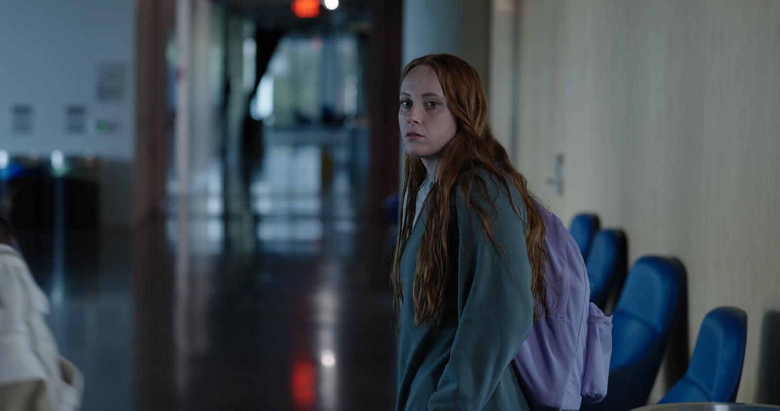 Young girl with long hair and backpack sitting on a row of chairs in a hallway.