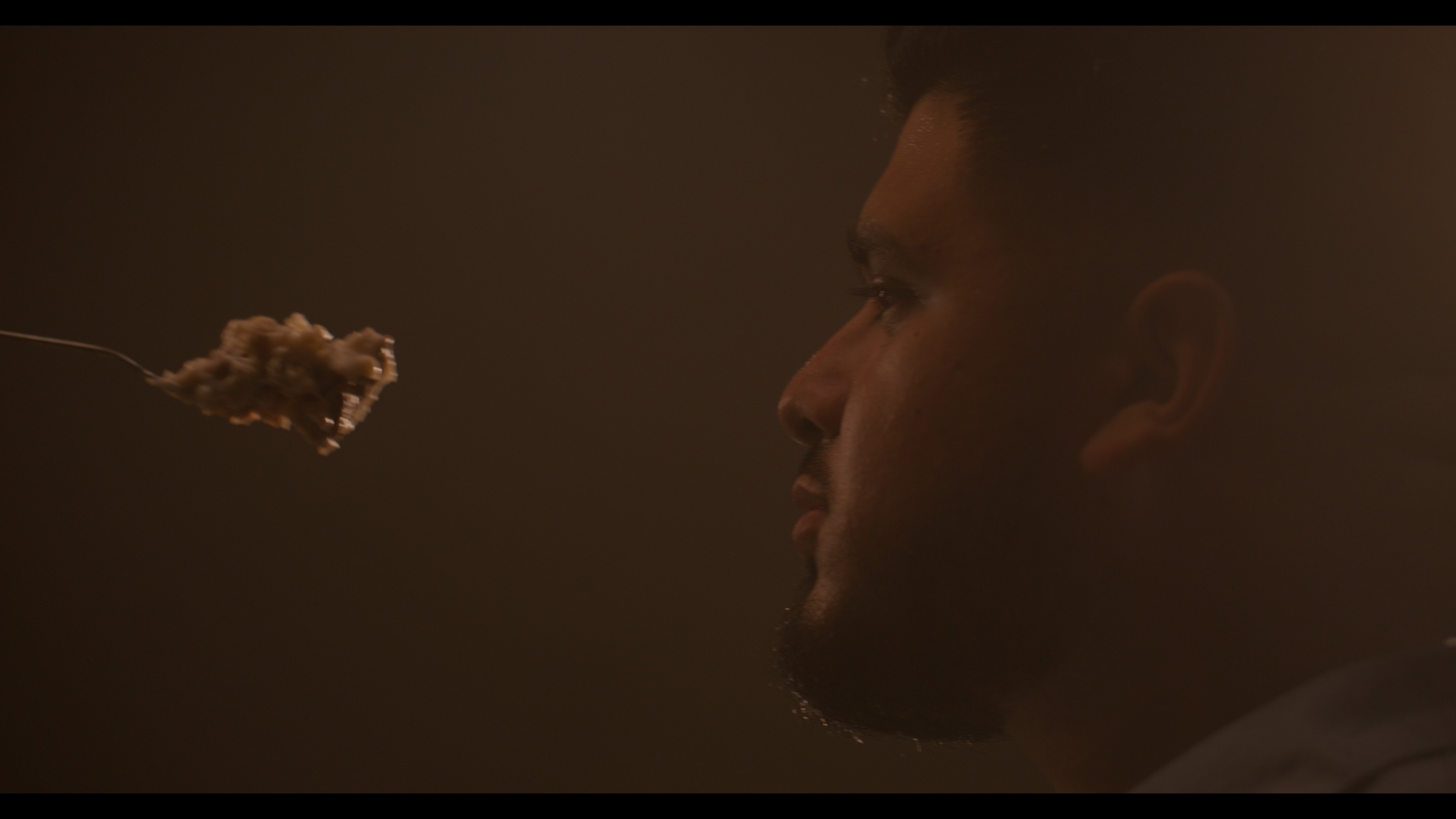 Side profile of a man facing a piece of food on a fork in a dimly lit setting.