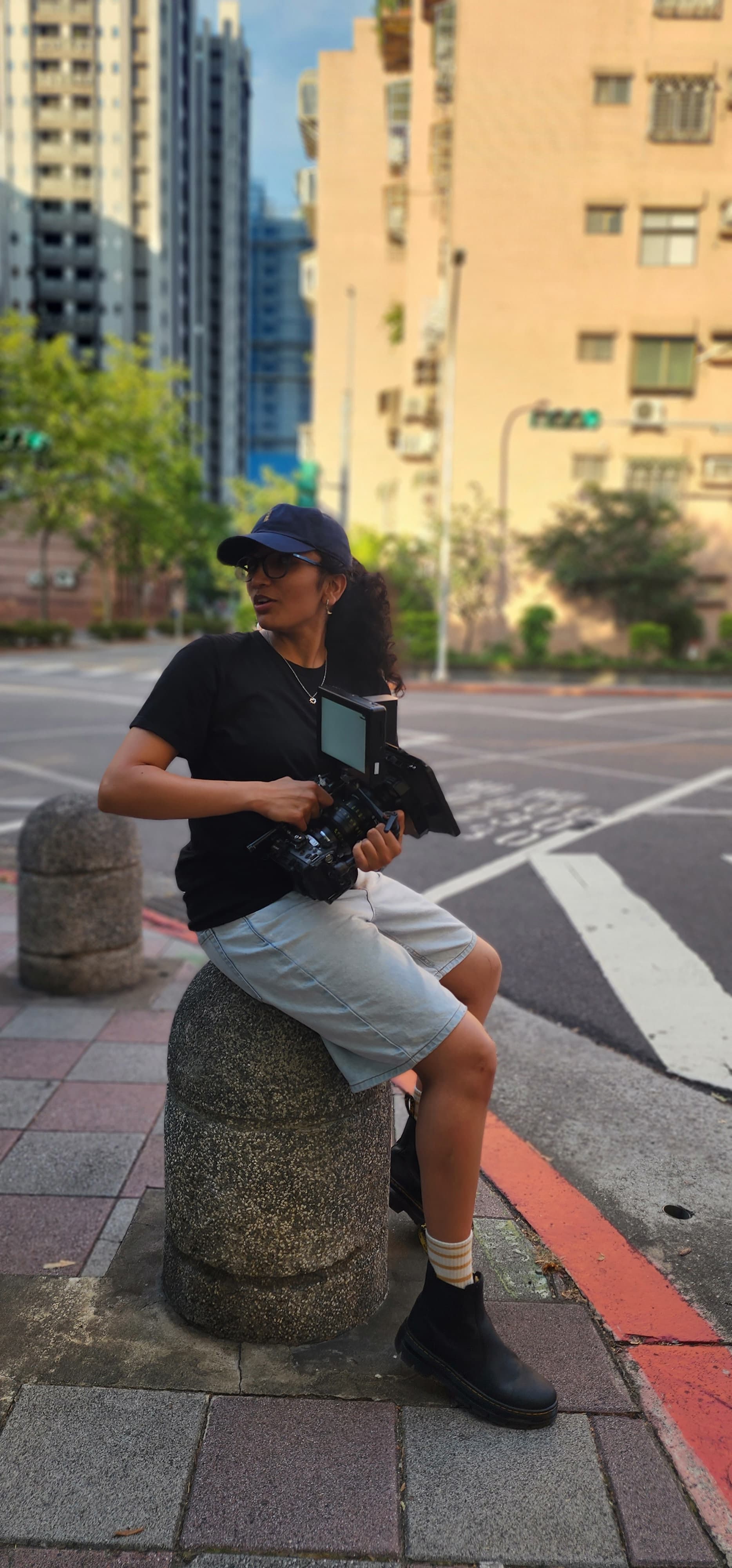 Person sitting on a concrete bollard holding a camera, wearing a black cap, black shirt, and gray shorts, in an urban setting.