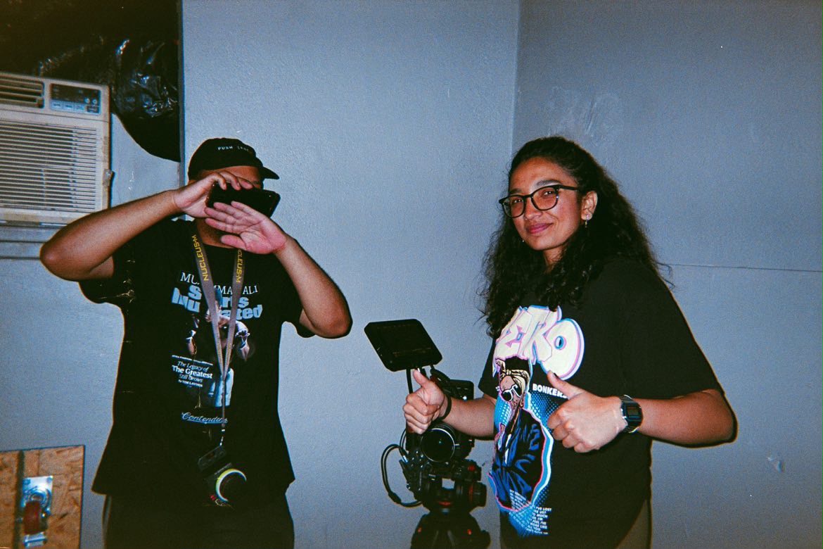 Two young women stand indoors, one covering her face with her hand, the other smiling and holding a camera, wearing glasses.