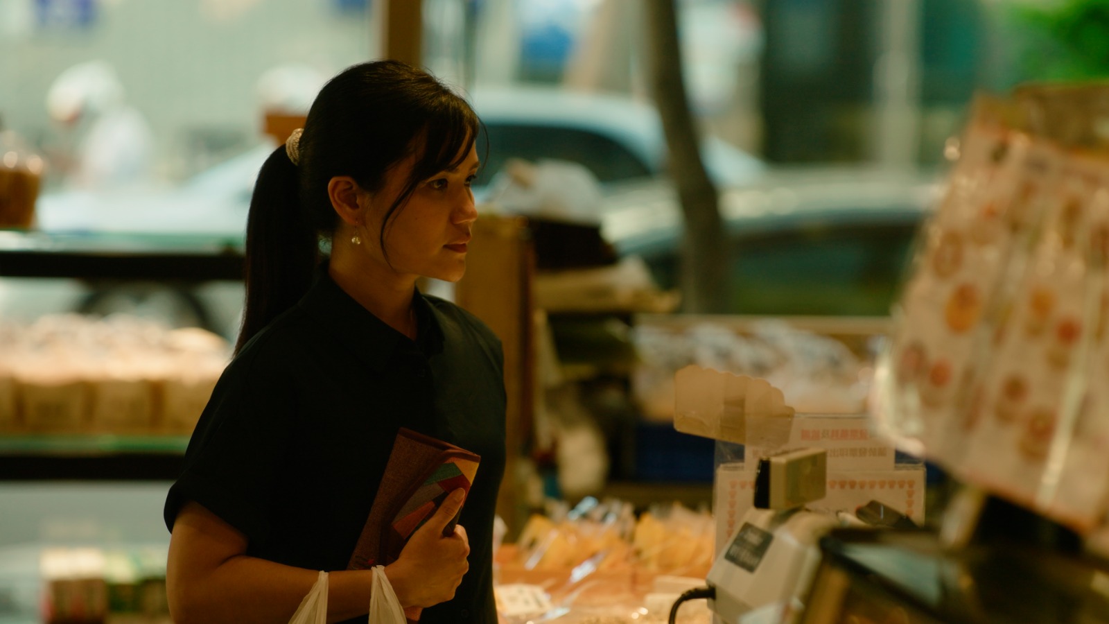 Young woman with dark hair in a ponytail holding a phone in a store, looking to the side, with shelves behind her.